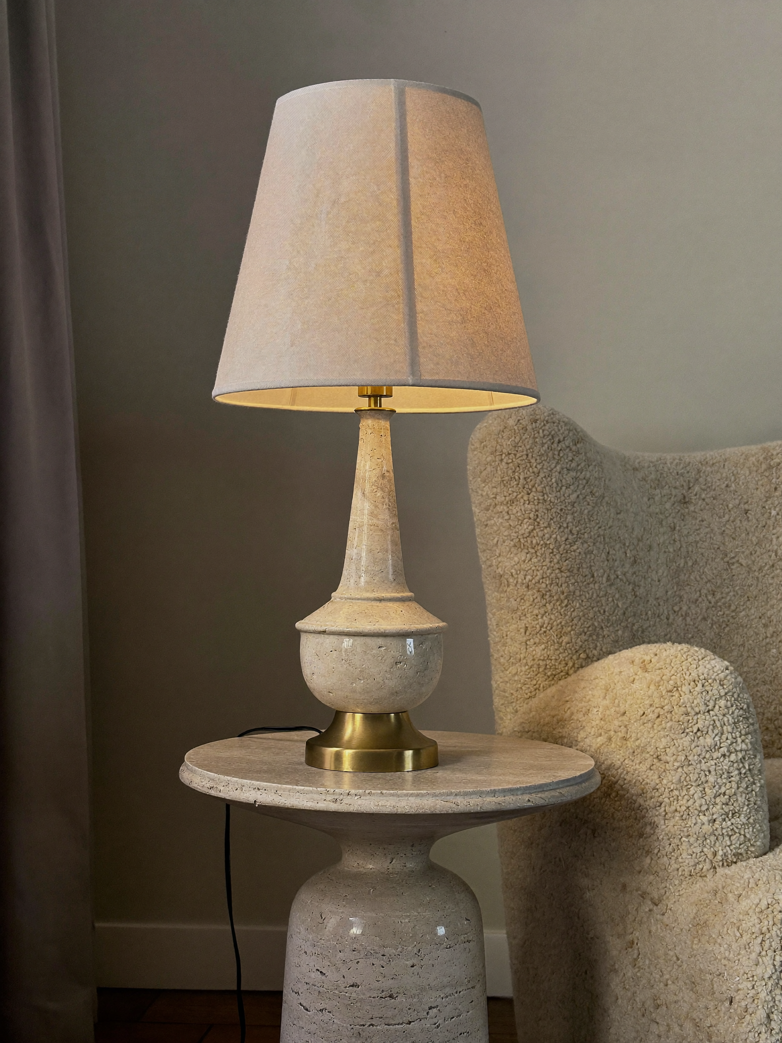 Linen and brass table lamp glowing on a travertine side table
